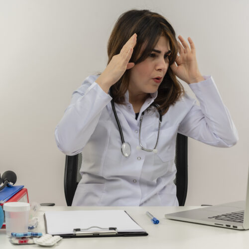 surprised middle-aged female doctor wearing medical robe and stethoscope sitting at desk with medical tools clipboard looking at laptop touching head isolated on white background